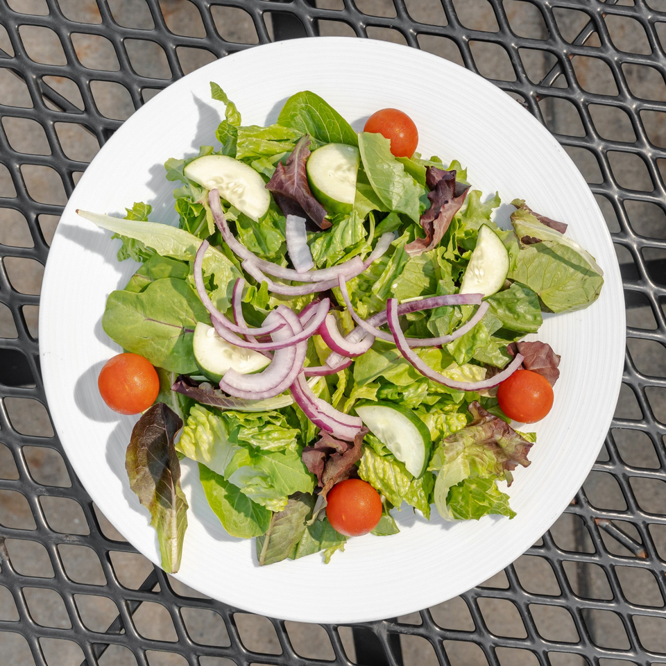 Fresh mixed green salad with cucumber slices, cherry tomatoes, and red onion on a white plate atop an outdoor metal table.