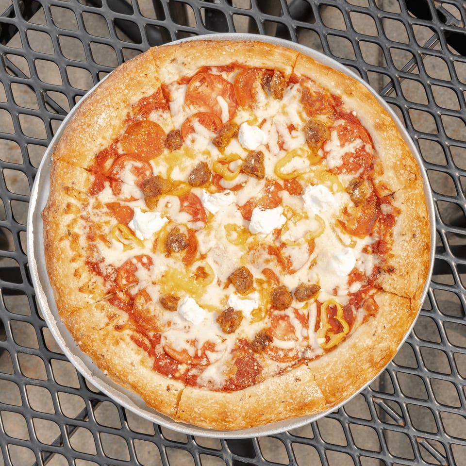 Overhead view of a golden-crusted pizza topped with melted cheese, pepperoni, sausage, tomatoes, and peppers on a metal pan.