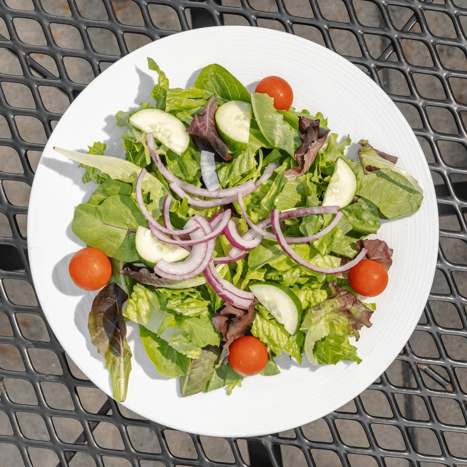 Fresh garden salad with mixed greens, cucumber slices, red onion, and cherry tomatoes on a white plate outdoors.