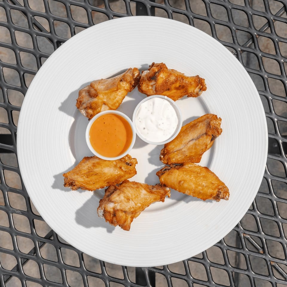 Six crispy chicken wings arranged on a white plate with orange buffalo sauce and creamy ranch dip on an outdoor metal table.