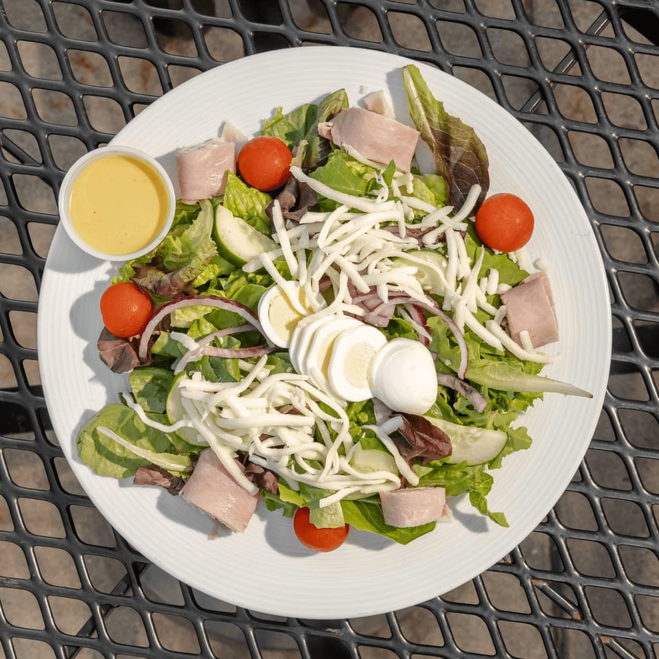 Garden salad with mixed greens, ham, tomatoes, cucumber, shredded cheese, red onion, boiled egg, and dressing on an outdoor table.
