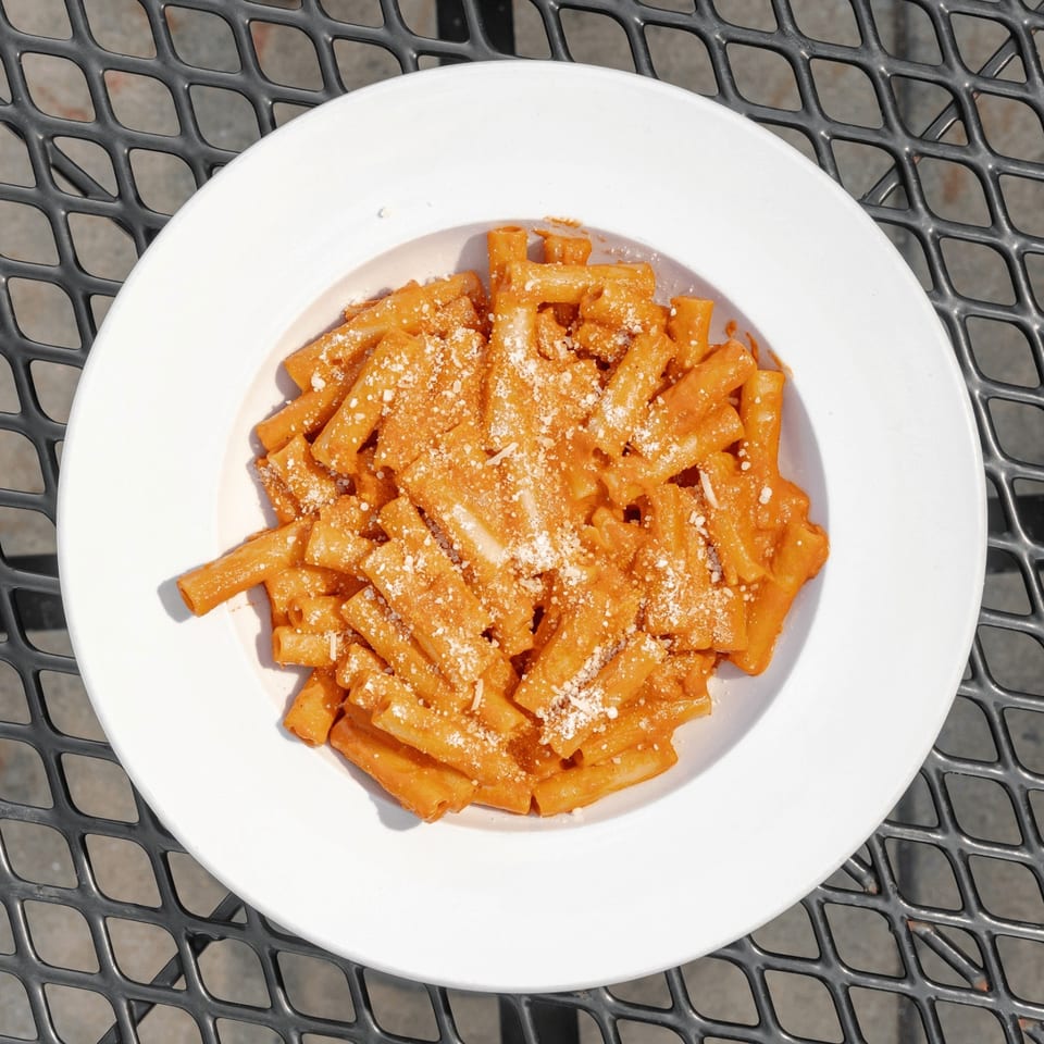 Rigatoni pasta coated in orange sauce and sprinkled with grated cheese on a white plate atop a black metal table.