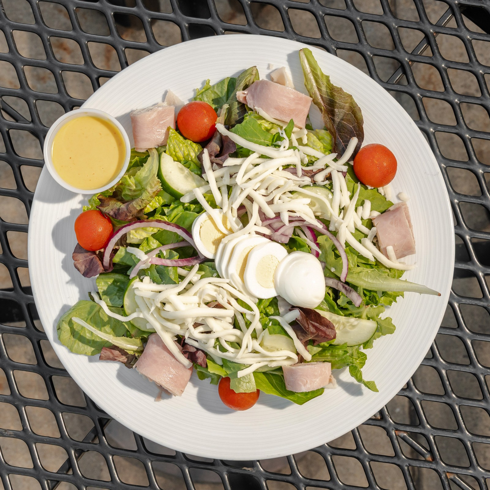 Garden salad with mixed greens, ham, hard-boiled egg, shredded cheese, tomatoes, cucumbers, onions, and dressing on a white plate.